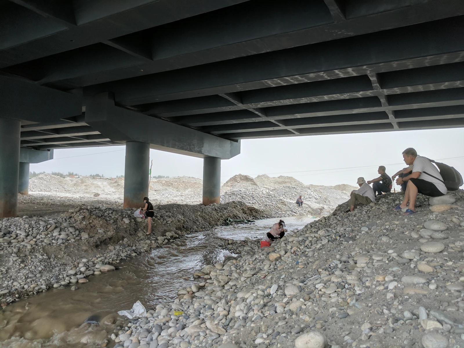 Jade collectors beneath the Yurungkash River bridge, 2017. The riverbed, the people reading it and a piece of modern infrastructure occupy the same frame. Photograph: Voidvector, CC BY-SA 4.0.